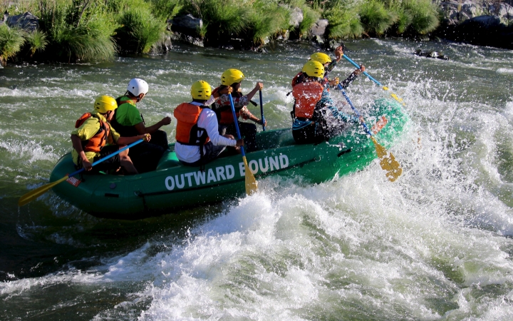 a group of outward bound students wearing helmets and life jackets paddle a raft through whitewater